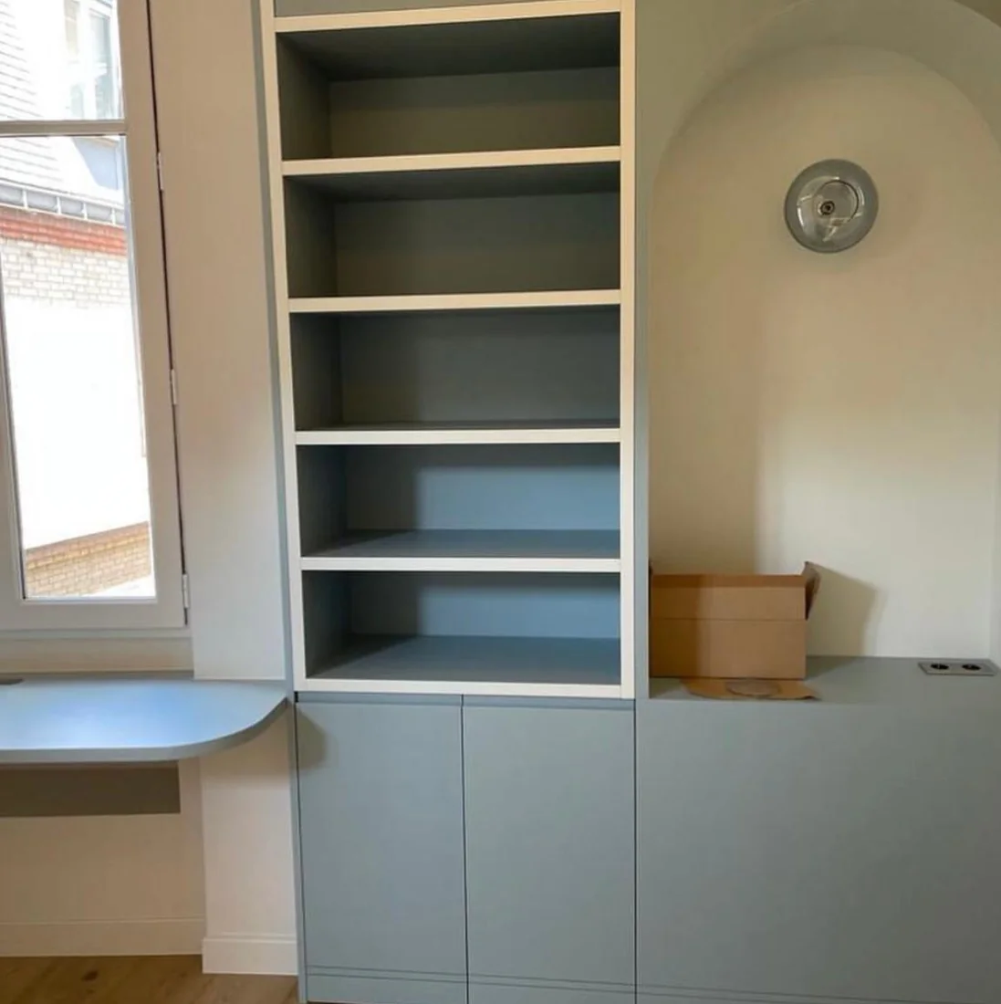 Rangement bleu-gris avec étagères ouvertes et tablette de bureau intégrée sous la fenêtre